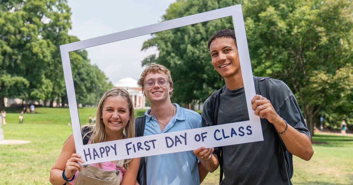 Three students outside, pose while holding a large Polaroid photo prop frame with the words happy first day of class written at the bottom.