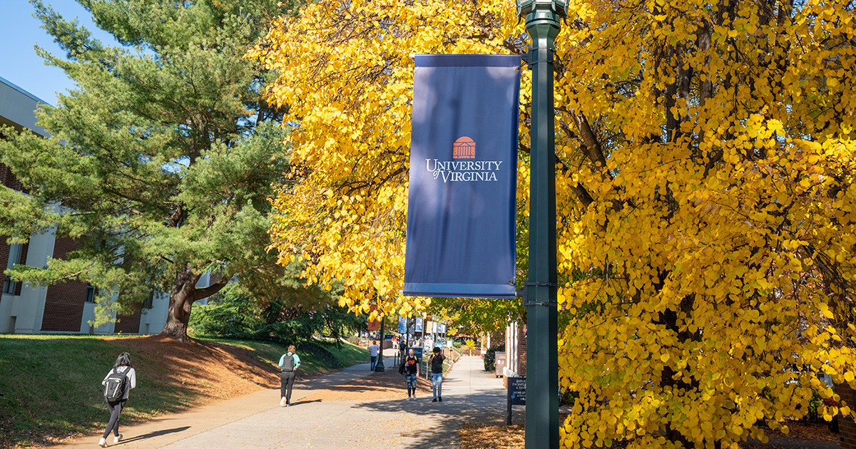 UVA dark blue banner is attached to a street light stand in front of golden fall leaves while students walk by.