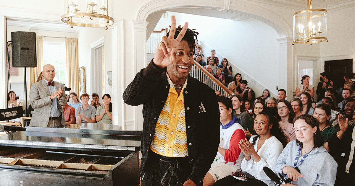 Musician Jon Batiste sits at a piano while talking to a group of students seated around him.